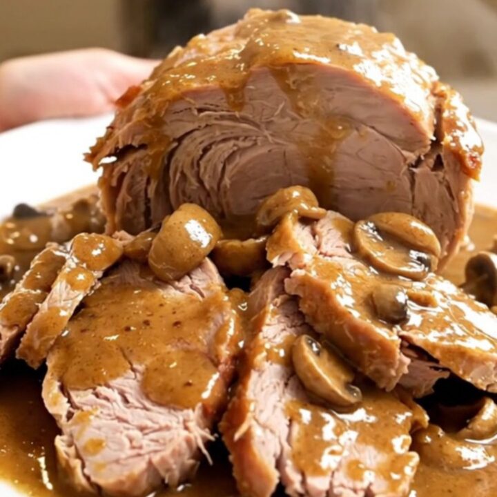 A slow-cooked pork roast with creamy mushroom sauce in a slow cooker on a white speckled countertop.