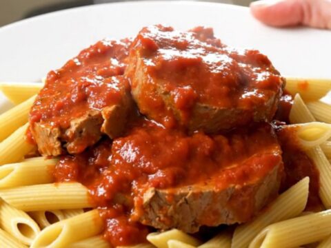 A top-down view of raw veal tendrons, sliced onions, and tomato coulis inside a black slow cooker insert.