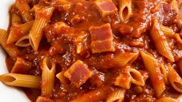A top-down view of a black slow cooker filled with penne pasta, bacon lardons, and a vibrant, spicy red tomato sauce.