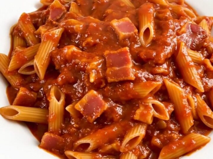 A top-down view of a black slow cooker filled with penne pasta, bacon lardons, and a vibrant, spicy red tomato sauce.