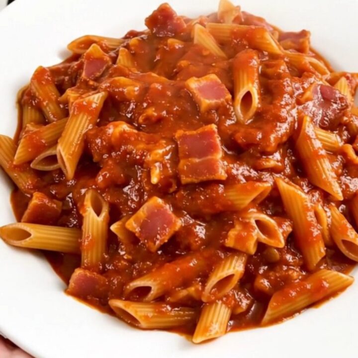 A top-down view of a black slow cooker filled with penne pasta, bacon lardons, and a vibrant, spicy red tomato sauce.
