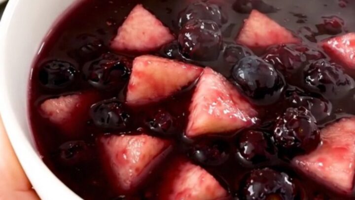 A close-up of a glossy, purple apple and wild blueberry compote in a white ceramic bowl.