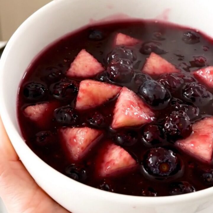 A close-up of a glossy, purple apple and wild blueberry compote in a white ceramic bowl.
