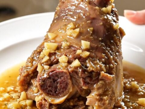 A close-up of a tender, glossy lamb shank on a white plate, coated in a rich garlic and rosemary sauce.