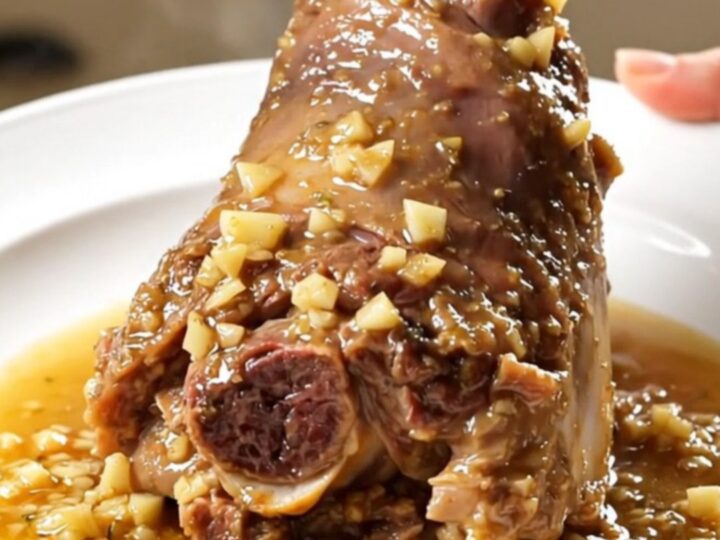A close-up of a tender, glossy lamb shank on a white plate, coated in a rich garlic and rosemary sauce.