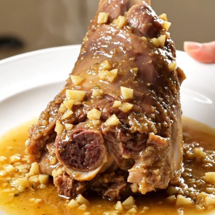 A close-up of a tender, glossy lamb shank on a white plate, coated in a rich garlic and rosemary sauce.