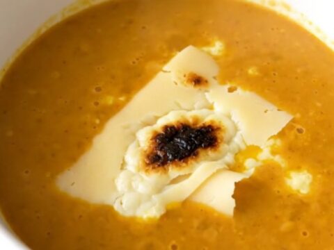A white bowl filled with creamy orange red kuri squash velouté topped with slices of melted Morbier cheese.