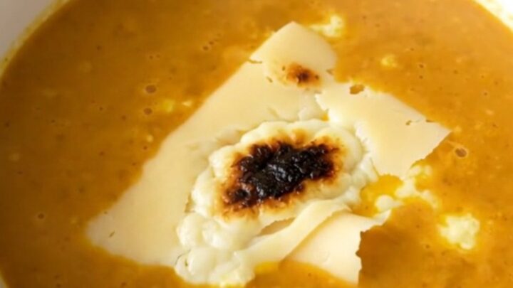 A white bowl filled with creamy orange red kuri squash velouté topped with slices of melted Morbier cheese.
