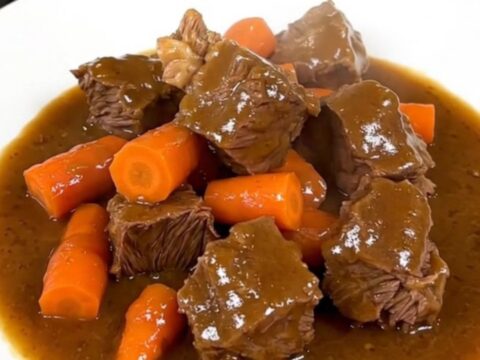 A top-down view of a black slow cooker filled with raw beef chunks and sliced carrots on a white quartz countertop.