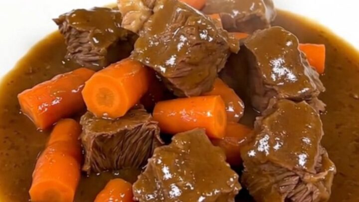 A top-down view of a black slow cooker filled with raw beef chunks and sliced carrots on a white quartz countertop.