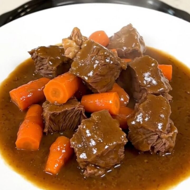 A top-down view of a black slow cooker filled with raw beef chunks and sliced carrots on a white quartz countertop.