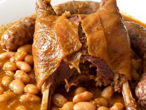 A rustic slow cooker filled with duck confit, sausages, and beans in a rich, glossy sauce.