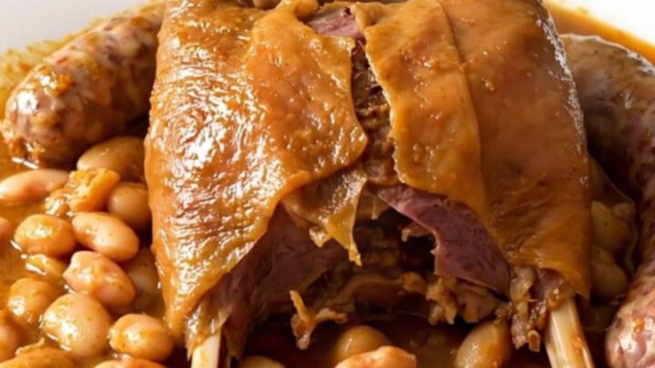 A rustic slow cooker filled with duck confit, sausages, and beans in a rich, glossy sauce.