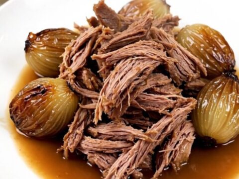 A close-up of tender, shredded braised lamb served with whole glossy shallots on a white plate.