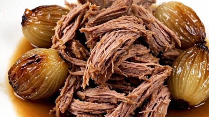 A close-up of tender, shredded braised lamb served with whole glossy shallots on a white plate.