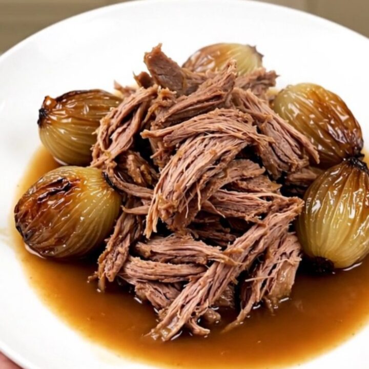 A close-up of tender, shredded braised lamb served with whole glossy shallots on a white plate.