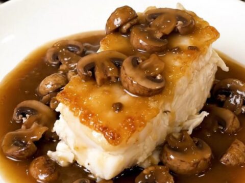 Tender white cod fillets topped with sliced mushrooms in a light cider broth served on a white plate.