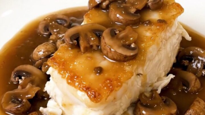 Tender white cod fillets topped with sliced mushrooms in a light cider broth served on a white plate.