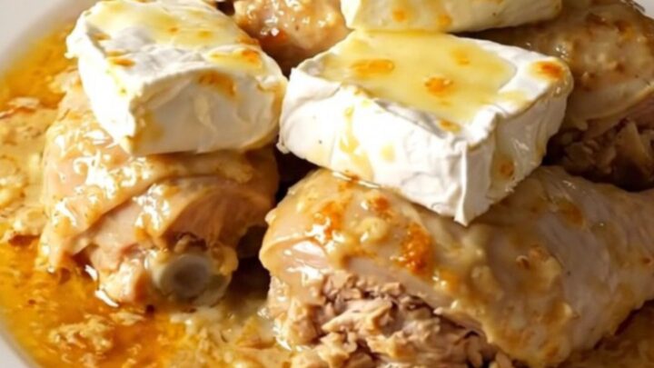 A top-down view of a black slow cooker filled with raw chicken, sliced onions, and slices of Camembert cheese.