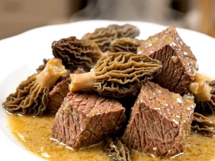 A close-up of tender braised beef cubes topped with morel mushrooms in a glossy, rich brown sauce on a white plate.