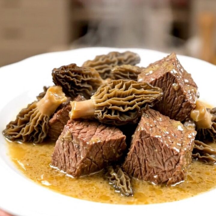 A close-up of tender braised beef cubes topped with morel mushrooms in a glossy, rich brown sauce on a white plate.