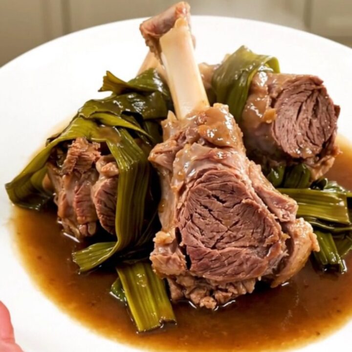 A top-down view of raw lamb chunks and sliced leeks in a black slow cooker on a white quartz countertop.