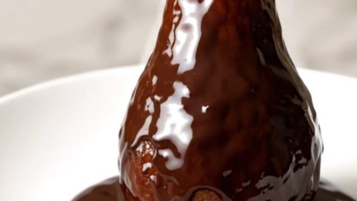 Poires entières pochées au chocolat noir brillant sur une assiette blanche.