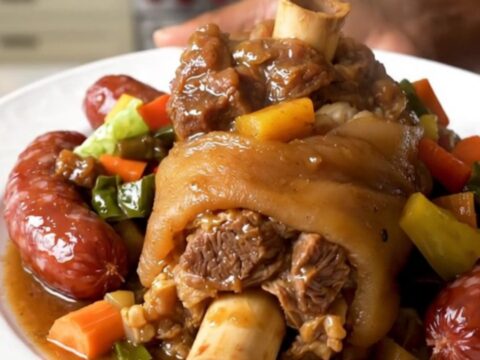 A rustic slow cooker filled with a hearty stew of pork shank, smoked sausages, cabbage, and root vegetables.