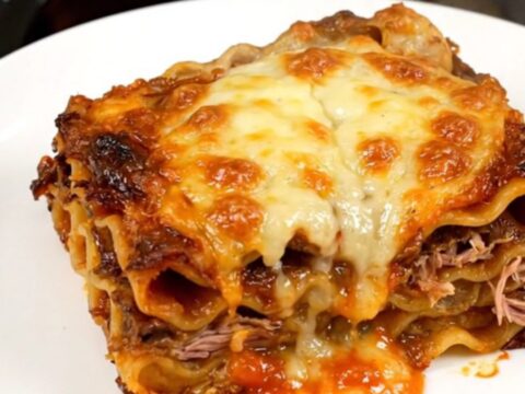 A top-down view of a black slow cooker filled with layers of lasagna, duck confit, mushrooms, and herbs.