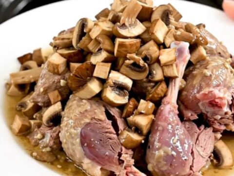 A close-up of slow-cooked guinea fowl with porcini mushrooms in a glossy white wine sauce on a white plate.