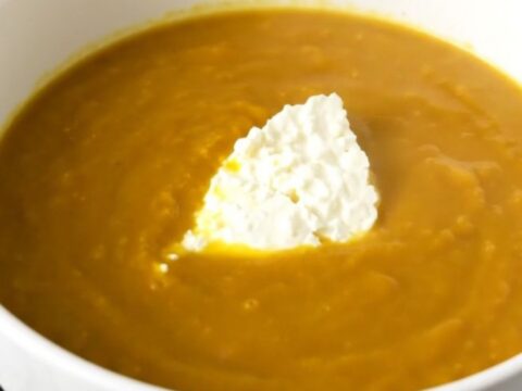 A white bowl filled with creamy orange pumpkin soup, topped with fresh goat cheese, sitting on a wooden table.