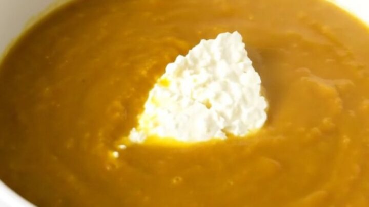 A white bowl filled with creamy orange pumpkin soup, topped with fresh goat cheese, sitting on a wooden table.