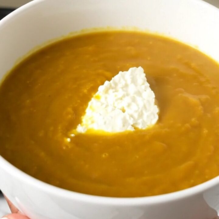 A white bowl filled with creamy orange pumpkin soup, topped with fresh goat cheese, sitting on a wooden table.