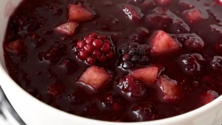 Glossy purple blackberry and apple compote served in a white bowl with a blurred kitchen background.
