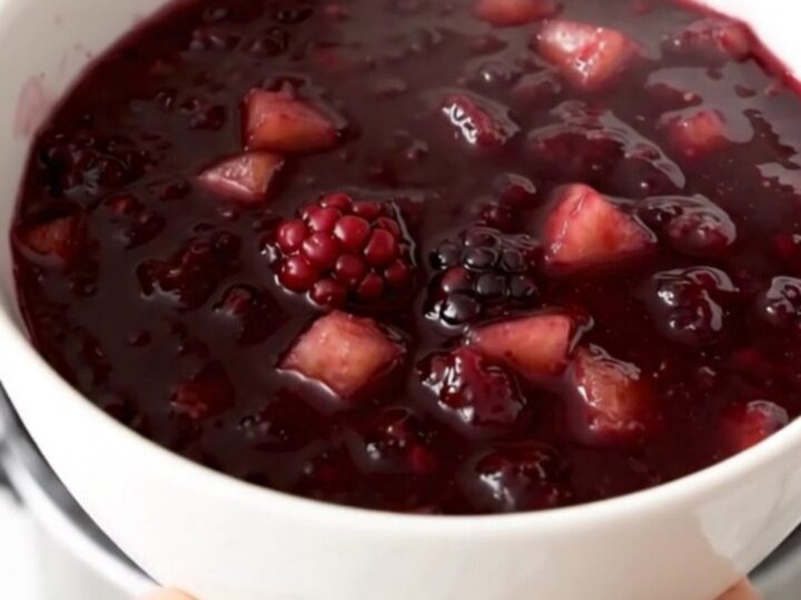 Glossy purple blackberry and apple compote served in a white bowl with a blurred kitchen background.