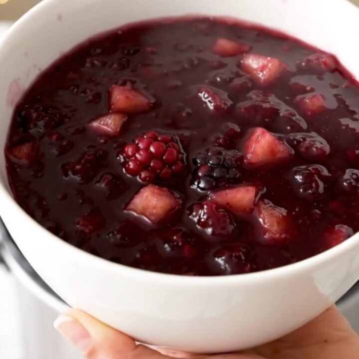 Glossy purple blackberry and apple compote served in a white bowl with a blurred kitchen background.