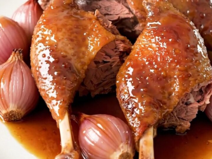 Close-up of slow-cooked, glossy honey balsamic duck legs served on a white plate with tender shallots.