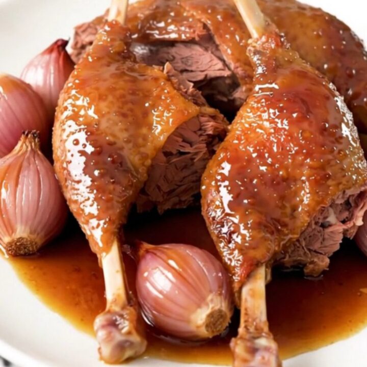 Close-up of slow-cooked, glossy honey balsamic duck legs served on a white plate with tender shallots.