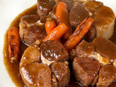 A close-up of tender pork cheeks in a glossy dark gravy with sliced carrots in a slow cooker.