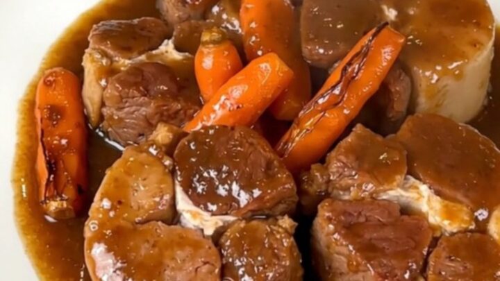 A close-up of tender pork cheeks in a glossy dark gravy with sliced carrots in a slow cooker.