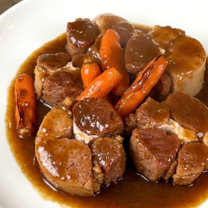A close-up of tender pork cheeks in a glossy dark gravy with sliced carrots in a slow cooker.