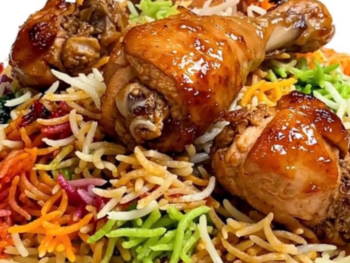 A steaming, glossy spiced chicken biryani served on a plain white plate held by a hand in a bright kitchen.