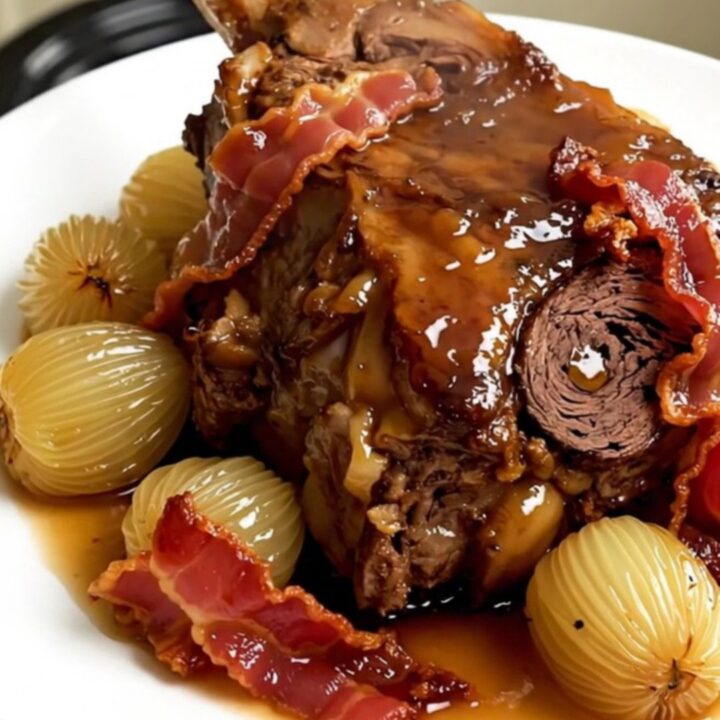 A rustic slow-cooked lamb shoulder with pearl onions and bacon on a white plate.