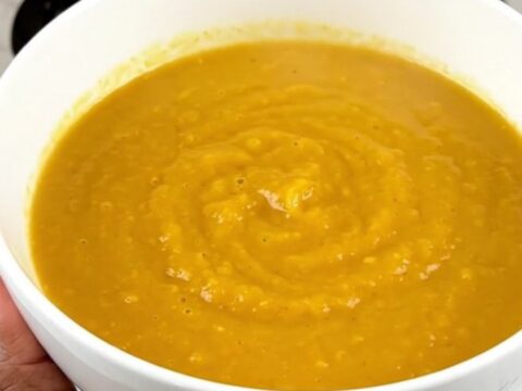 A steaming bowl of vibrant orange carrot, orange, and coriander soup on a white speckled quartz countertop.