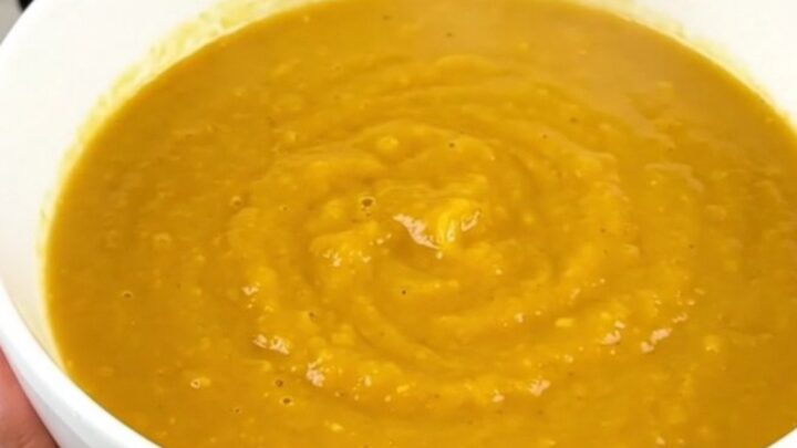 A steaming bowl of vibrant orange carrot, orange, and coriander soup on a white speckled quartz countertop.