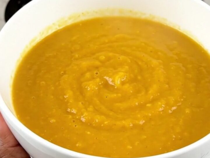 A steaming bowl of vibrant orange carrot, orange, and coriander soup on a white speckled quartz countertop.