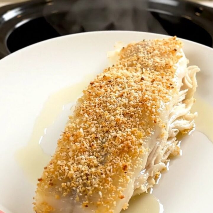 A top-down view of a black slow cooker on a white quartz countertop with raw pollock fillets, shallots, parsley