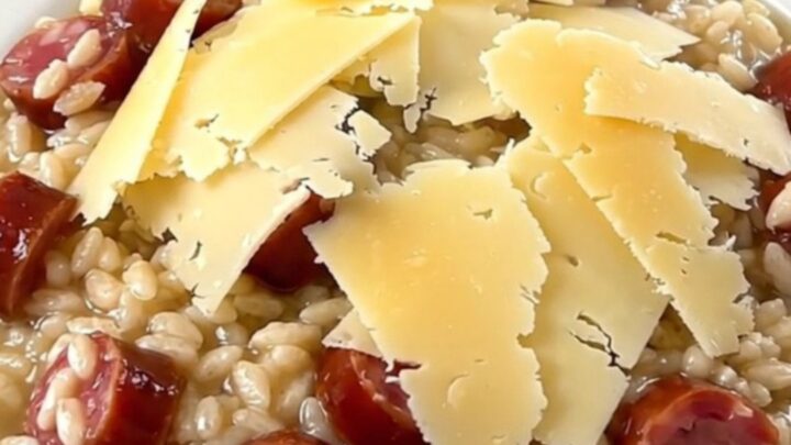 A close-up of a steaming, glossy bowl of risotto topped with melted Comté cheese and sliced sausage.