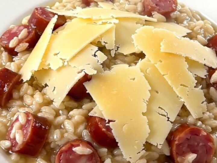 A close-up of a steaming, glossy bowl of risotto topped with melted Comté cheese and sliced sausage.