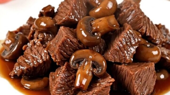 A close-up of tender, glossy braised beef chunks served with mushrooms on a white plate.